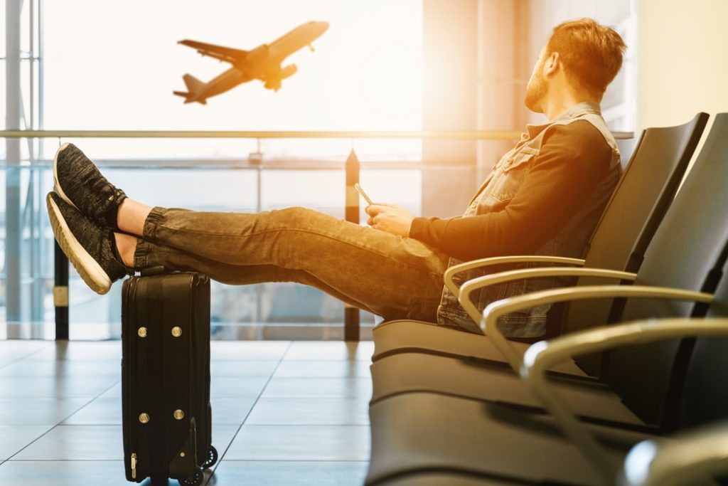 Un chico sentado en el aeropuerto con sus pies sobre la maleta mira por el cristal cómo un avión despega.